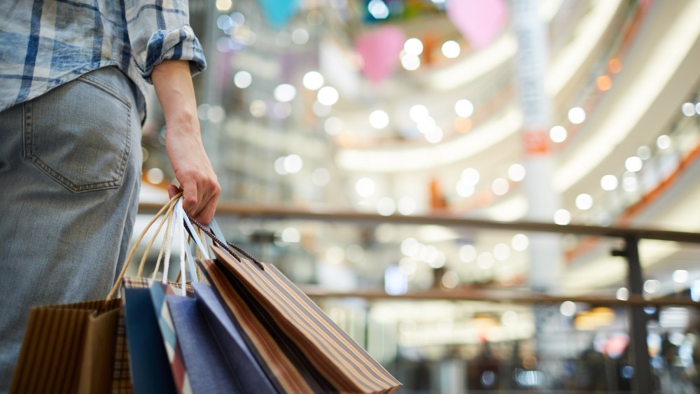 Close-up of unrecognizable woman in casual clothing standing in spacious shopping mall with lights and holding many paper bags