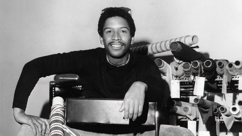 Jay Jaxon, photographed smiling in front of rolls of fabric at the Maison of Jean-Louis Scherrer’s design salon, to advertise the release of his 1970 Spring/Summer, Haute Couture & Prêt-à-Porter collections, 1969/1970