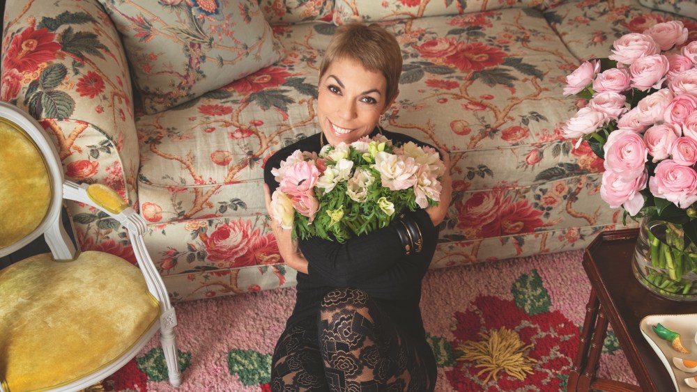 Mathilde Favier weating a lace bodysuit sitting on a floral carpet holding a bouquet of fresh flowers.