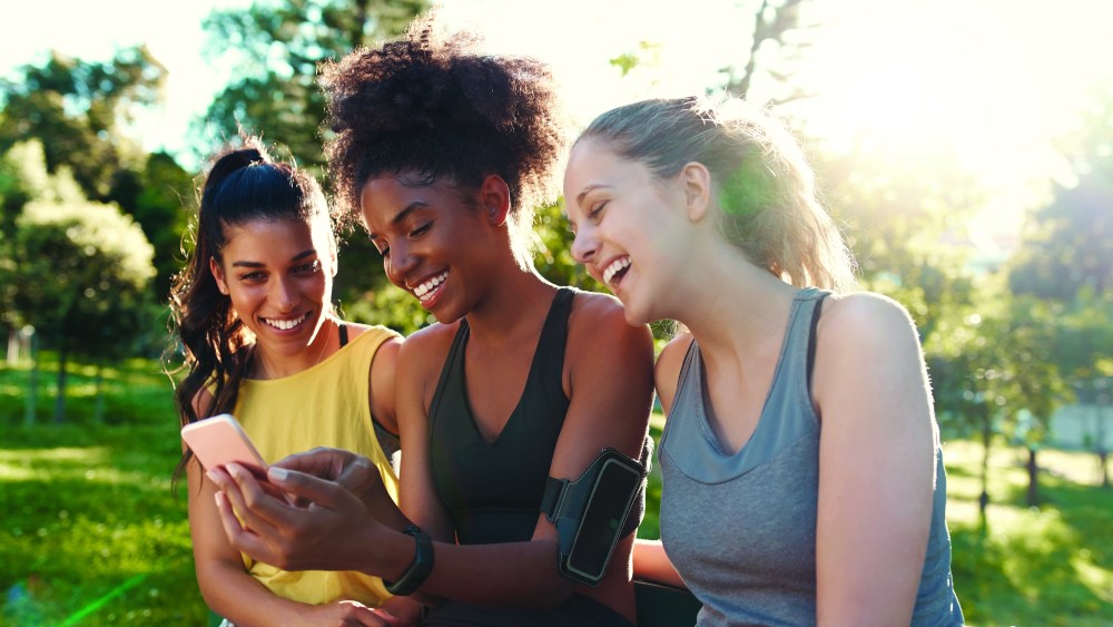 Fit young woman showing something on mobile phone to her friends - friends laughing together